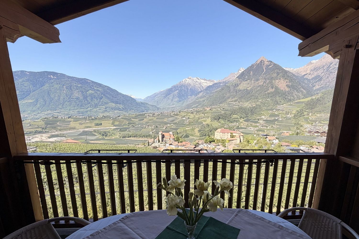 Ein Blick von einem hölzernen Balkon auf ein Tal mit Weinbergen, einem kleinen Ort mit Kirche und Schloss, umrahmt von hohen Alpenbergen bei klarem Himmel.