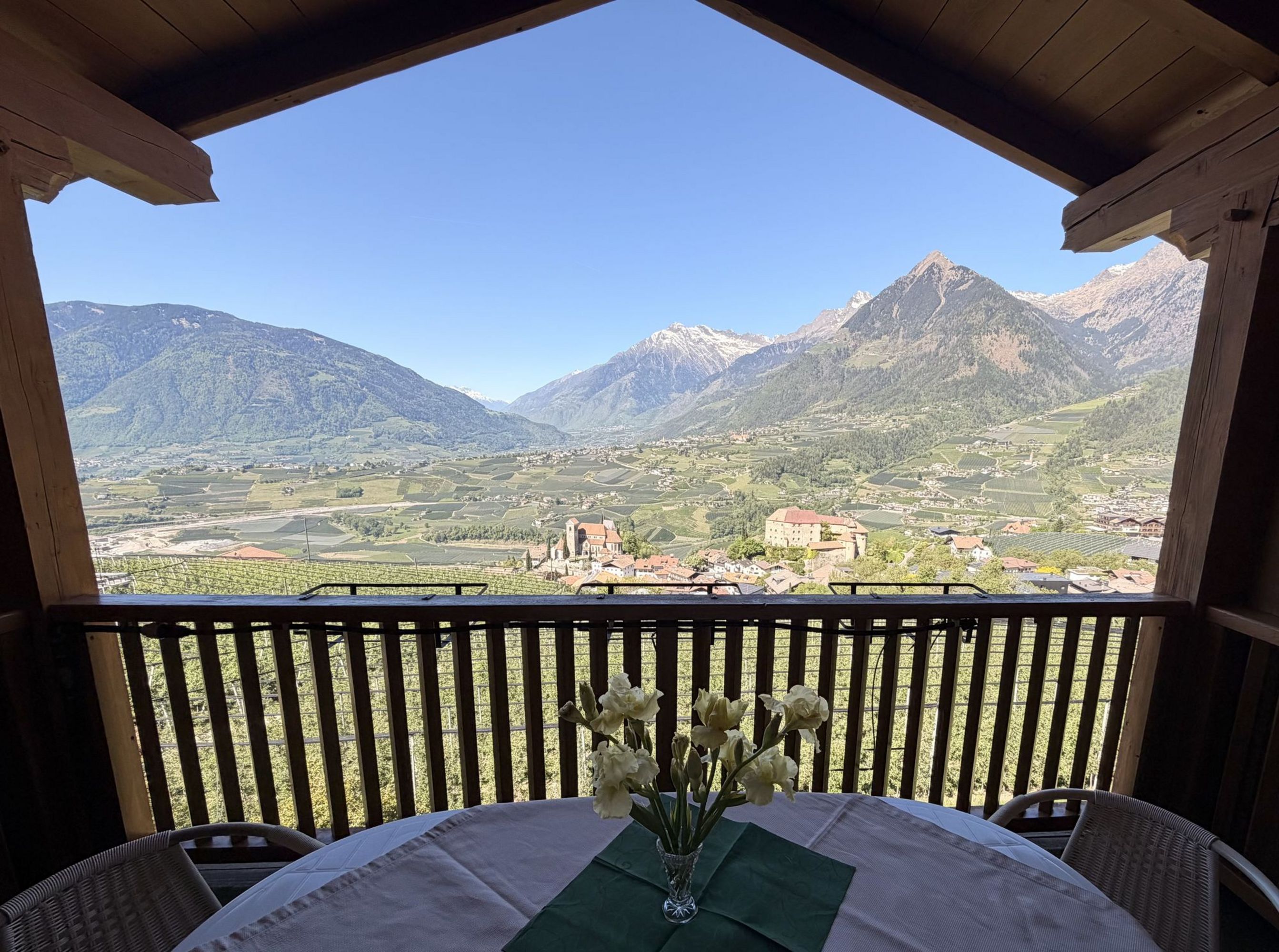 Ein Blick von einem hölzernen Balkon auf ein Tal mit Weinbergen, einem kleinen Ort mit Kirche und Schloss, umrahmt von hohen Alpenbergen bei klarem Himmel.