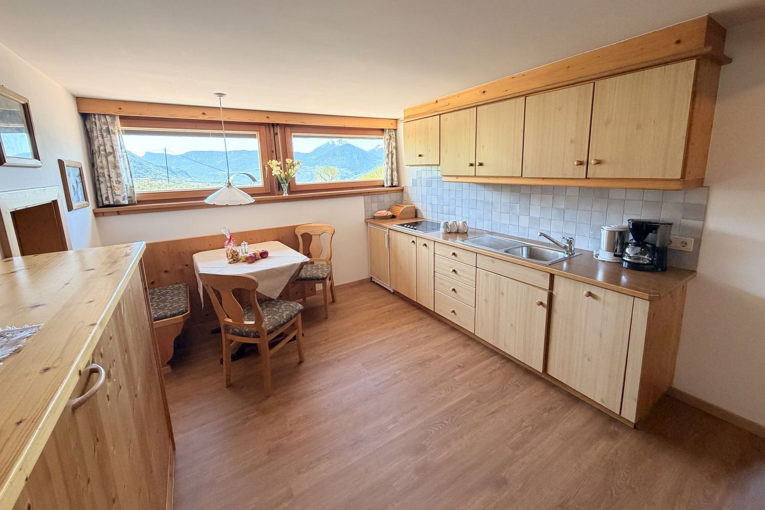 Eine rustikale Küche mit hellen Holzmöbeln, einer Sitzecke mit Tisch und zwei Stühlen sowie einem Fenster mit Bergblick.