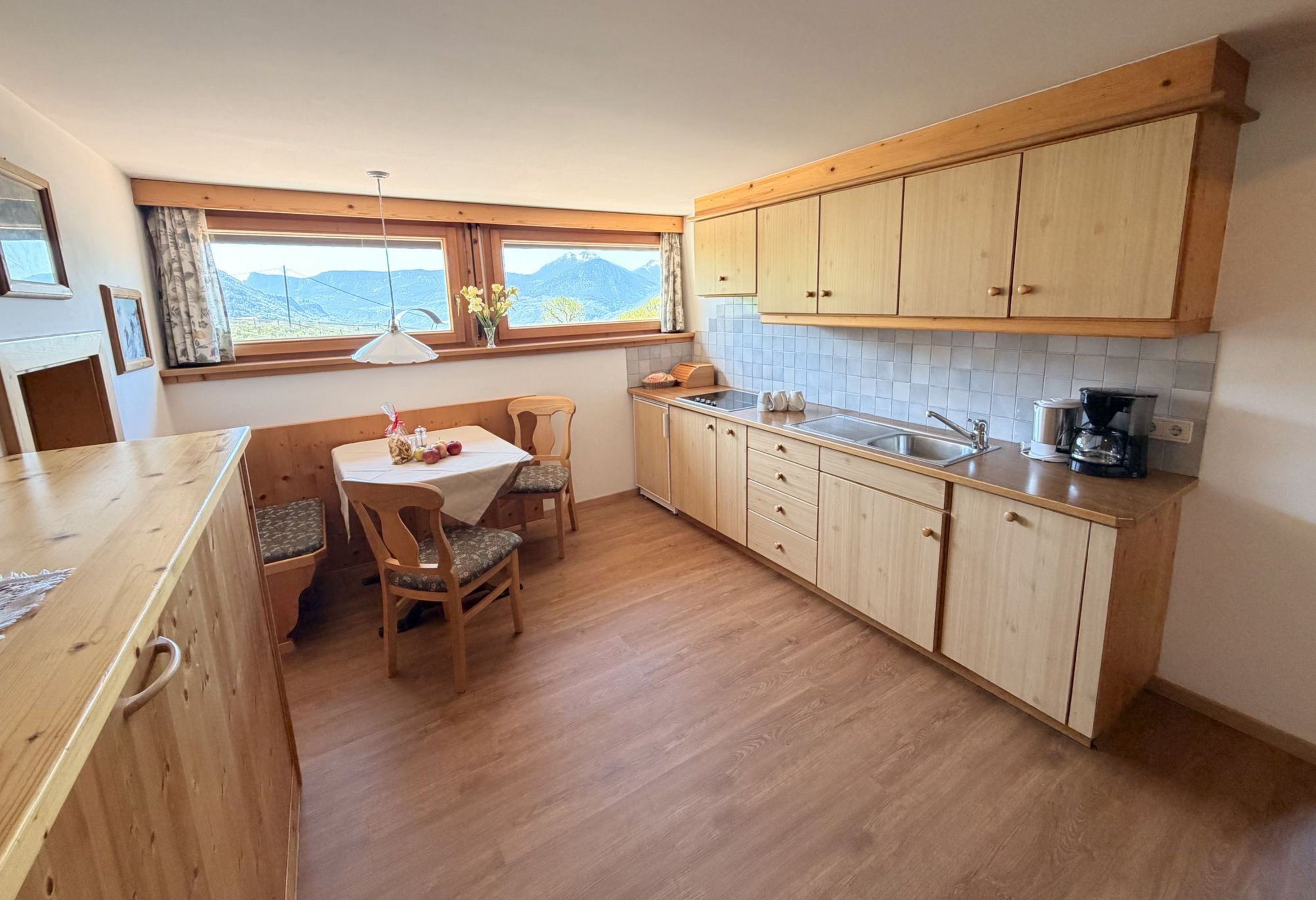 Eine rustikale Küche mit hellen Holzmöbeln, einer Sitzecke mit Tisch und zwei Stühlen sowie einem Fenster mit Bergblick.