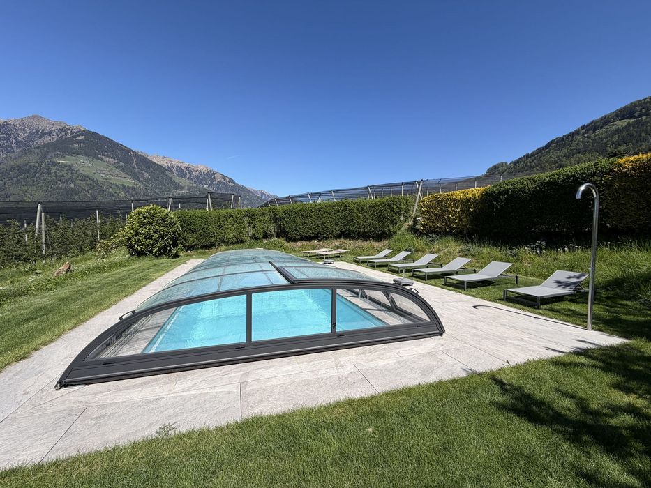 Ein Außenpool mit gläserner Überdachung befindet sich auf einer gepflegten Rasenfläche, umgeben von Sonnenliegen und im Hintergrund erheben sich steile Berge unter einem klaren blauen Himmel.