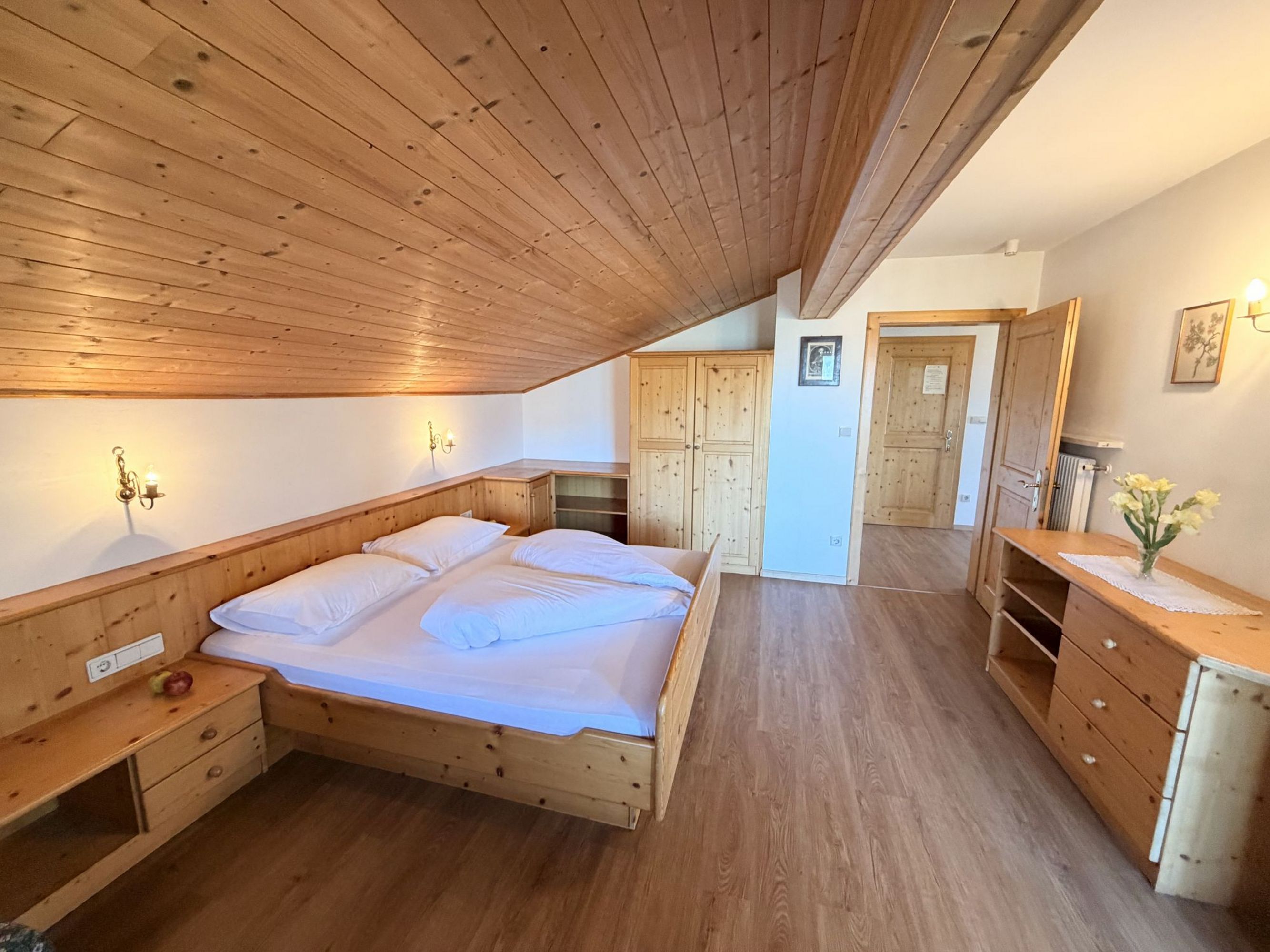 Ein gemütliches Schlafzimmer unter einer beigen Holzschräge mit einem Holzbett, einem Kleiderschrank und einer Kommode vor einer offenen Tür.