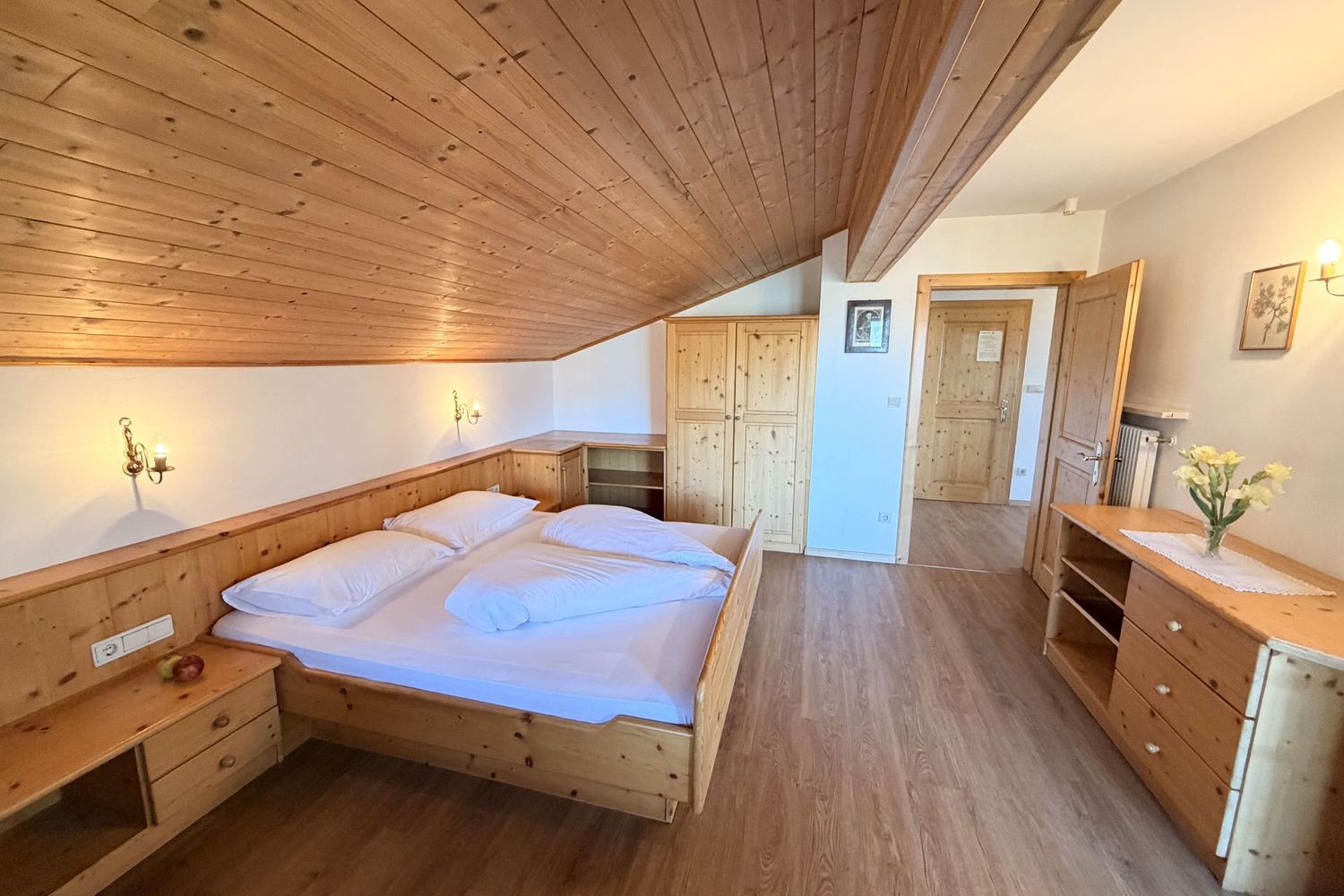 Ein gemütliches Schlafzimmer unter einer beigen Holzschräge mit einem Holzbett, einem Kleiderschrank und einer Kommode vor einer offenen Tür.