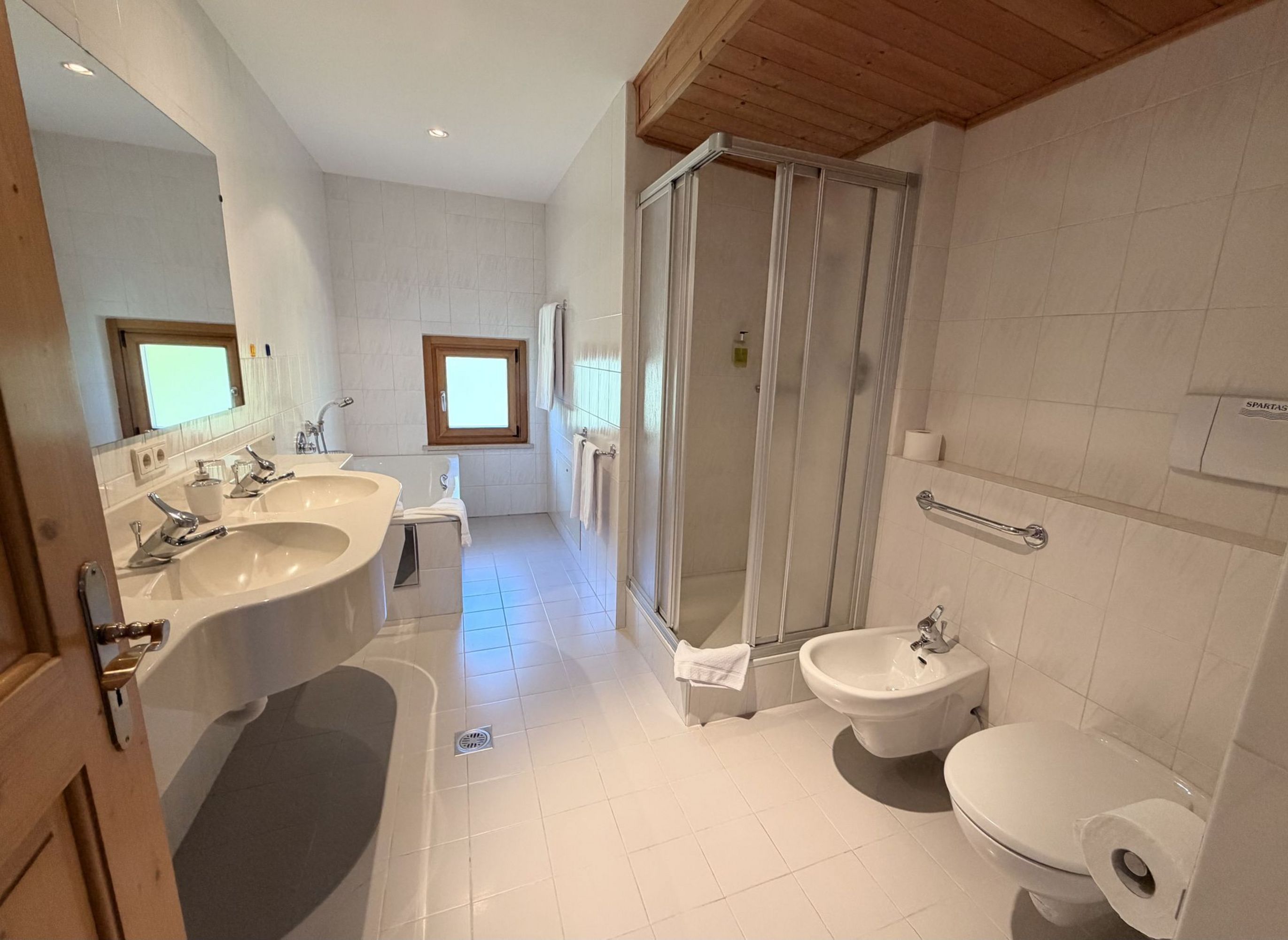 Ein helles, gefliestes Badezimmer mit Doppelwaschbecken, einer Duschkabine, einer Toilette und einem Bidet.