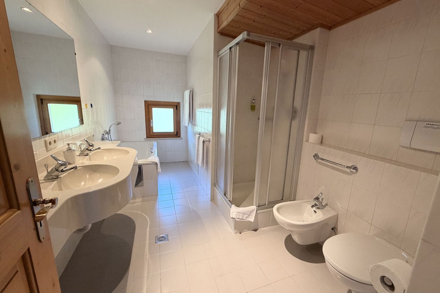 Ein helles, gefliestes Badezimmer mit Doppelwaschbecken, einer Duschkabine, einer Toilette und einem Bidet.