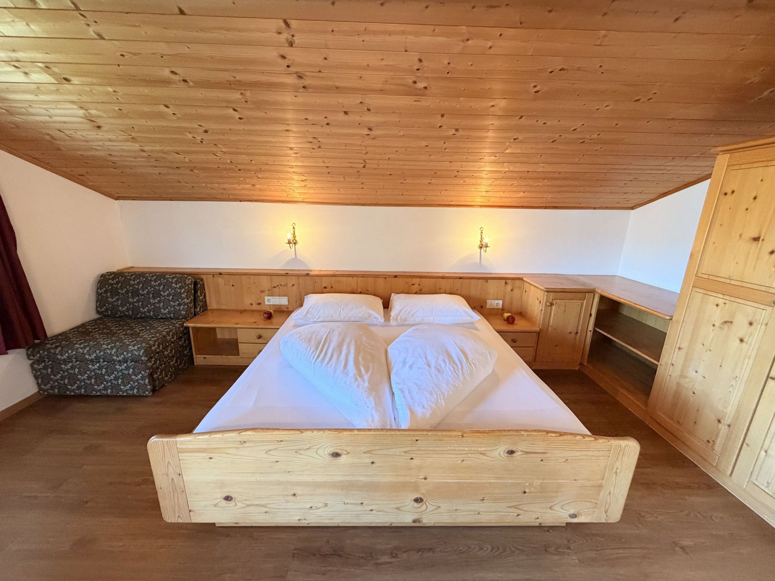 Ein Schlafzimmer unter einer Dachschräge mit einem Holzbett, weißen Bettdecken, einem gemusterten Sessel und Holzschränken.
