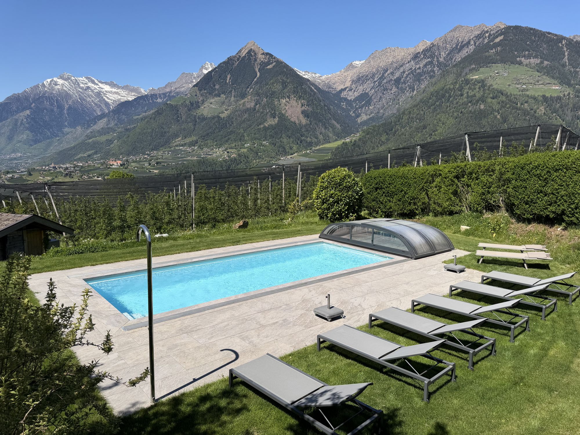 Ein rechteckiger Außenpool mit blauem Wasser und ausziehbarer Abdeckung steht umgeben von einer gefliesten Terrasse und Liegestühlen in einer bergigen Landschaft.